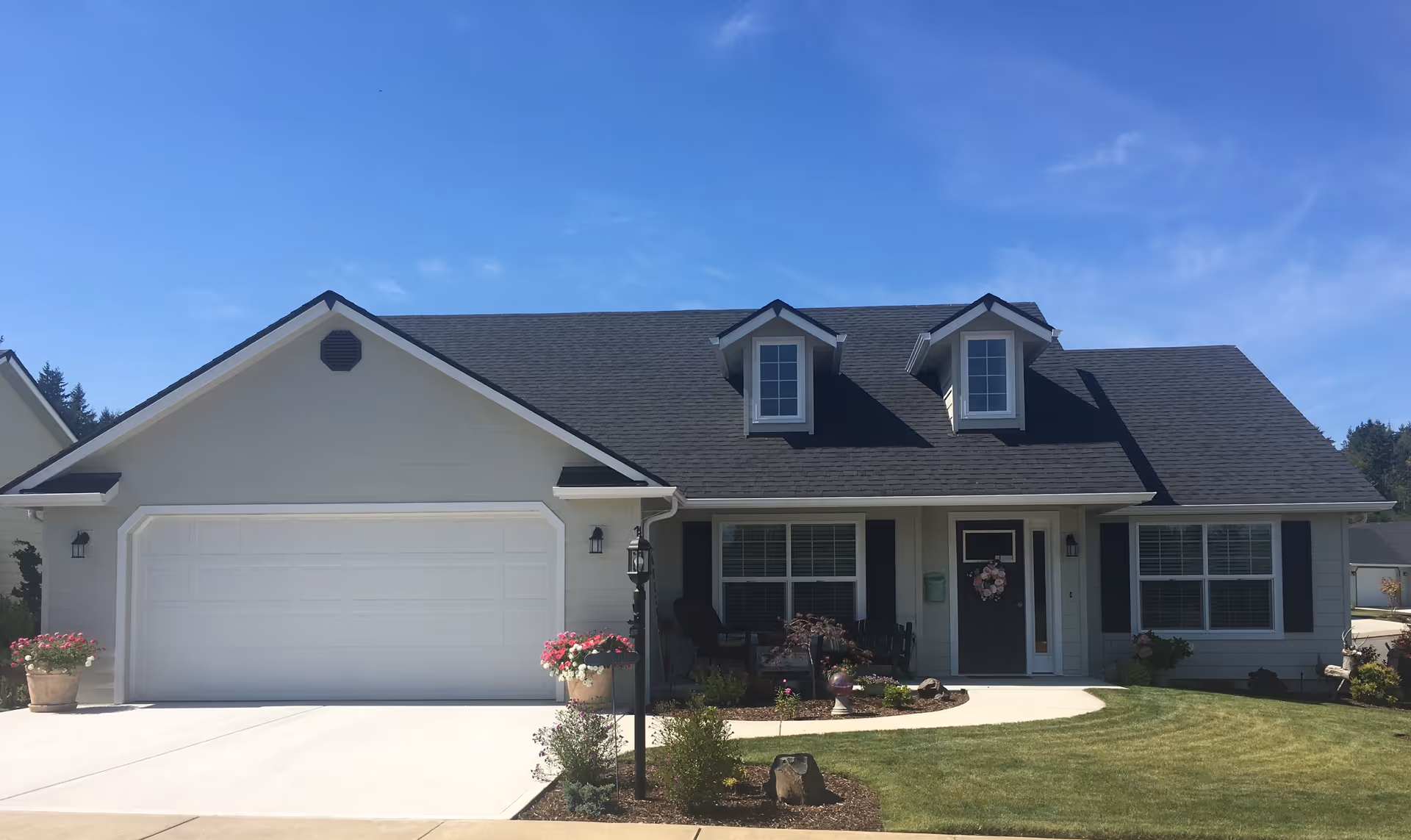 Front exterior view of a single-story house with a dark shingled roof, two dormer windows, a double garage door, a front porch with seating, and a well-maintained lawn with flower pots and landscaping under a clear blue sky.