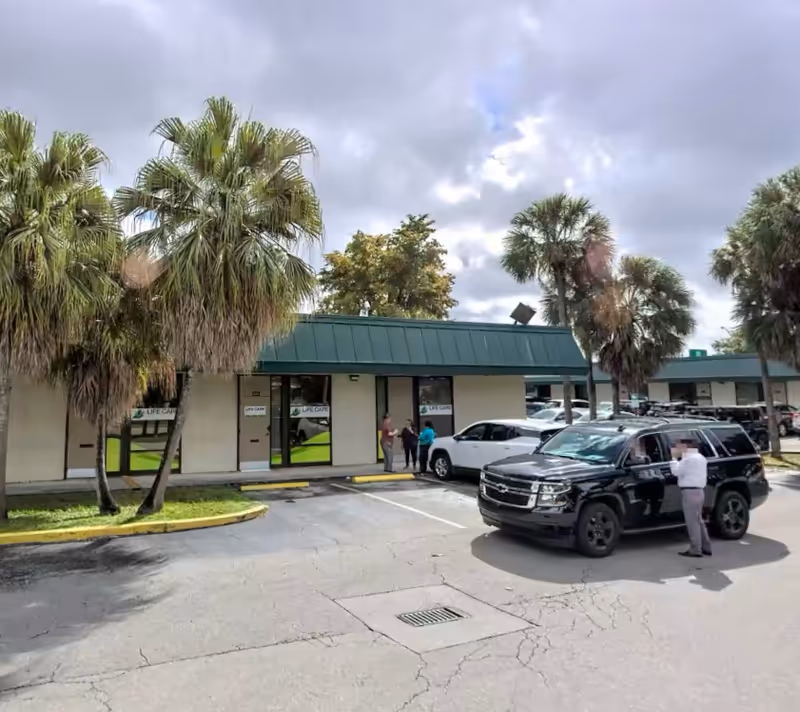 Exterior view of Simply Home Care Miami with storefront entrances, palm trees, and parked cars.