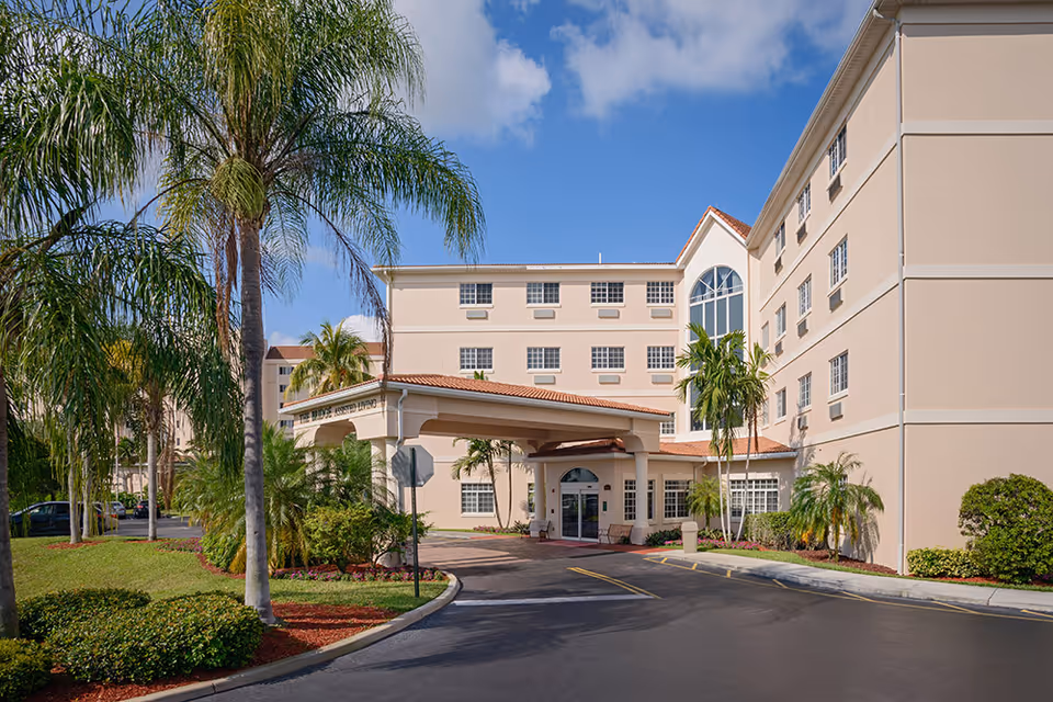 Exterior view of a multi-story senior living facility with beige walls and a red-tiled roof. The entrance features a covered drop-off area with palm trees and landscaped greenery surrounding the driveway under a blue sky with some clouds.