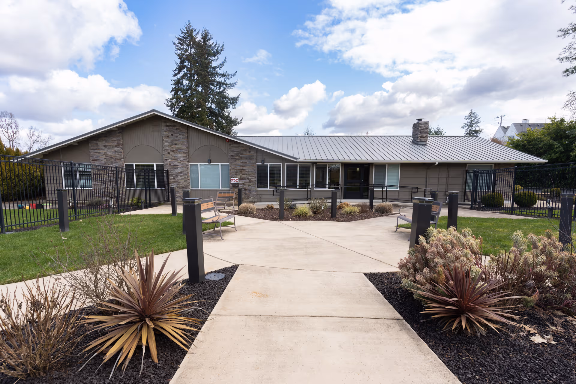 Exterior view of a single-story building with a metal roof and stone accents, surrounded by a fenced garden area with benches, plants, and a concrete walkway leading to the entrance under a partly cloudy sky.