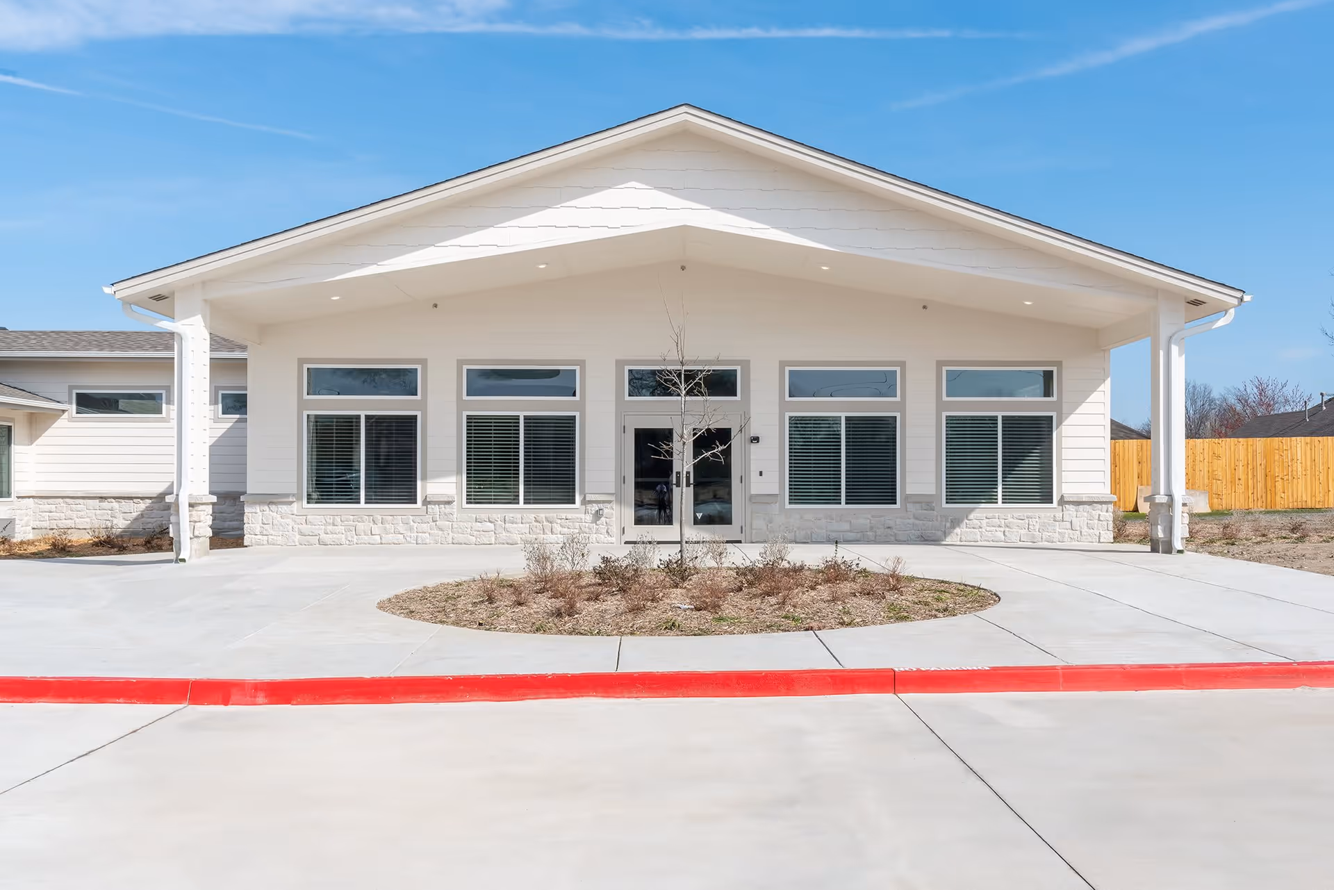 Front exterior view of a single-story building with a white facade, large windows with blinds, a central glass door, and a covered entrance. There is a circular landscaped area with a small tree and shrubs in front of the building, and a red curb along the driveway.