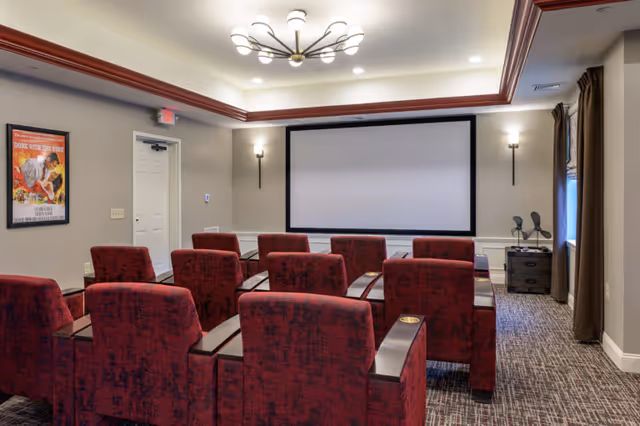 A small theater room with two rows of red upholstered chairs facing a large blank projection screen. The room has beige walls, a patterned carpet, a modern ceiling light fixture, wall sconces, a framed poster on the left wall, and a window with brown curtains on the right.
