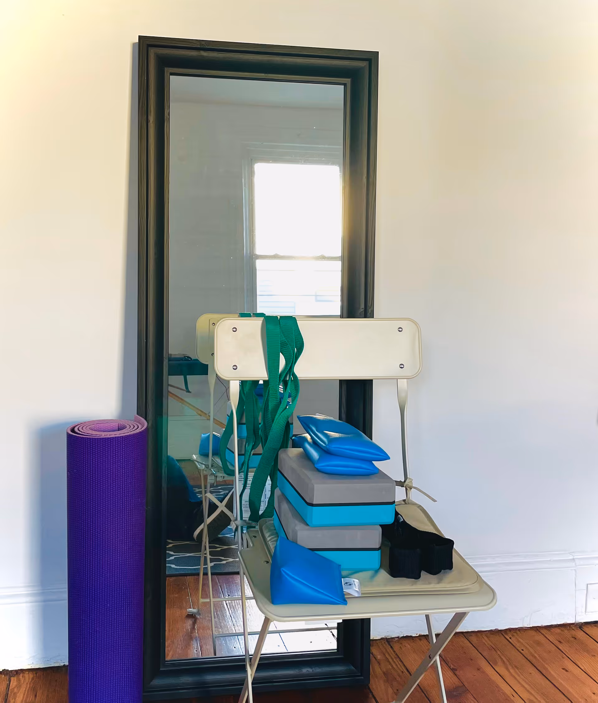 Full-length mirror reflecting exercise chair, stacked blocks, straps, and a purple yoga mat in a room.