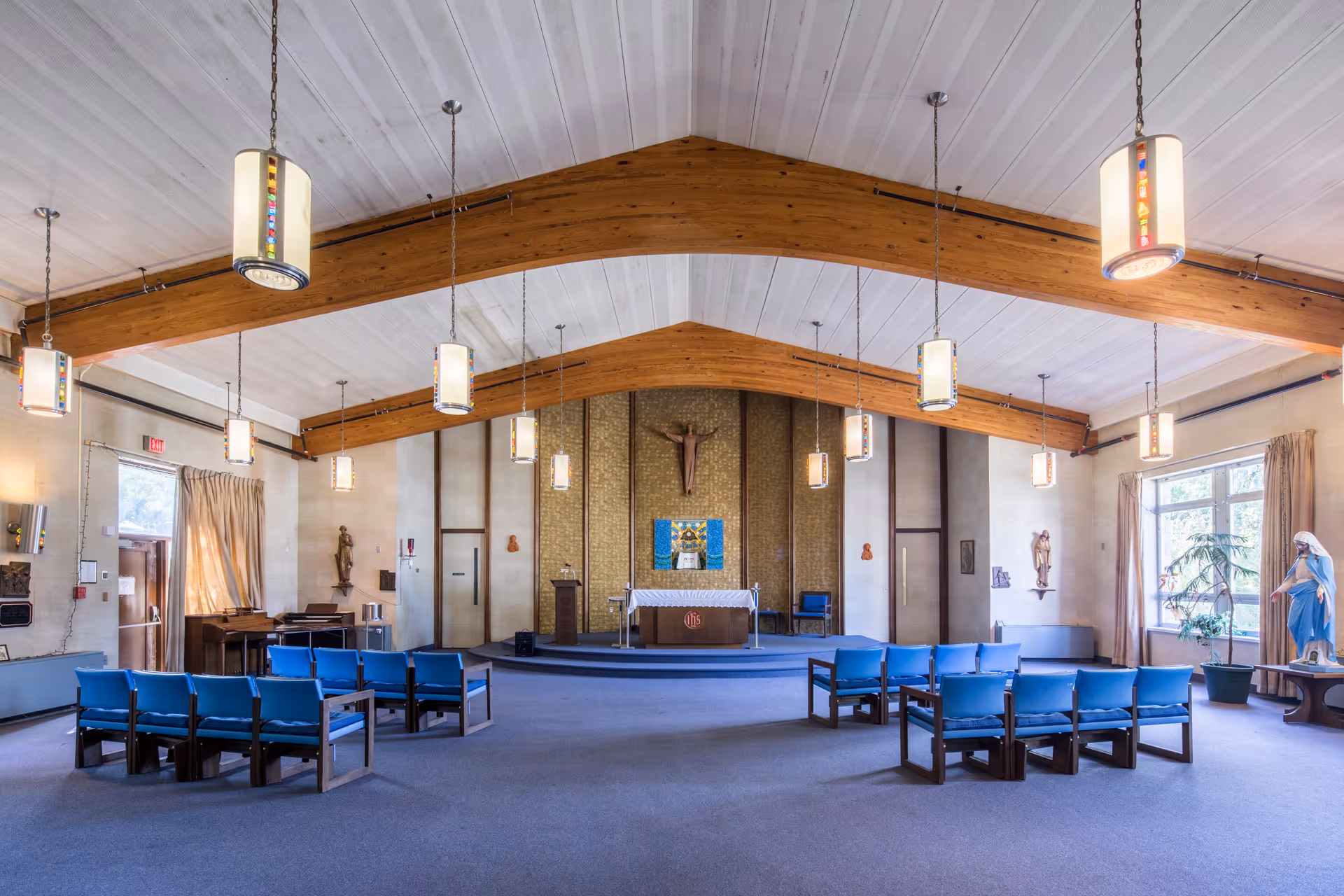 Interior view of a chapel or worship room with wooden beams on the ceiling, hanging pendant lights, blue cushioned chairs arranged in rows, religious statues, and an altar with a crucifix on the back wall.