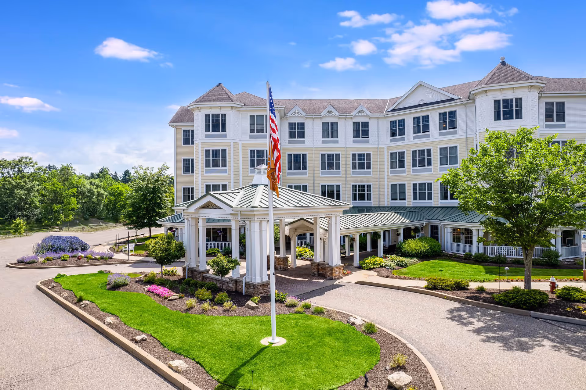 Exterior view of a multi-story senior living facility with a covered entrance, landscaped greenery, and an American flag on a flagpole under a blue sky with some clouds.