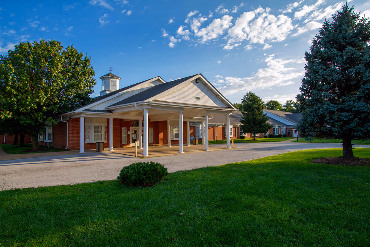 Front exterior view of Oakwood Estates Nursing & Rehab building with a covered entrance, surrounded by green grass, trees, and a partly cloudy blue sky.