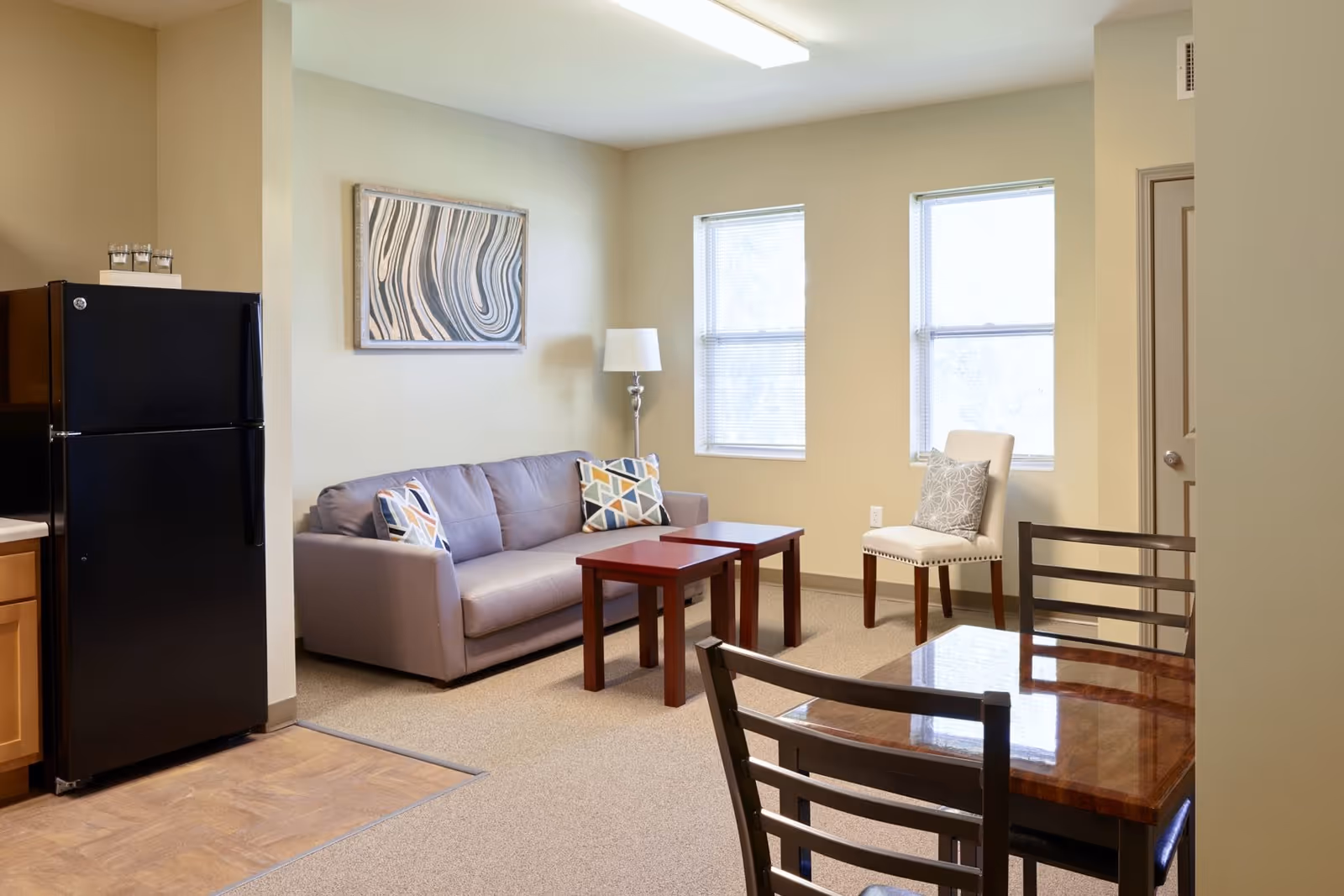 A cozy living area in a senior living facility featuring a gray sofa with patterned cushions, two small wooden coffee tables, a white upholstered chair with a decorative pillow, and a floor lamp. There are two windows with blinds letting in natural light. A black refrigerator and part of a kitchen counter are visible on the left, and a wooden dining table with chairs is in the foreground.