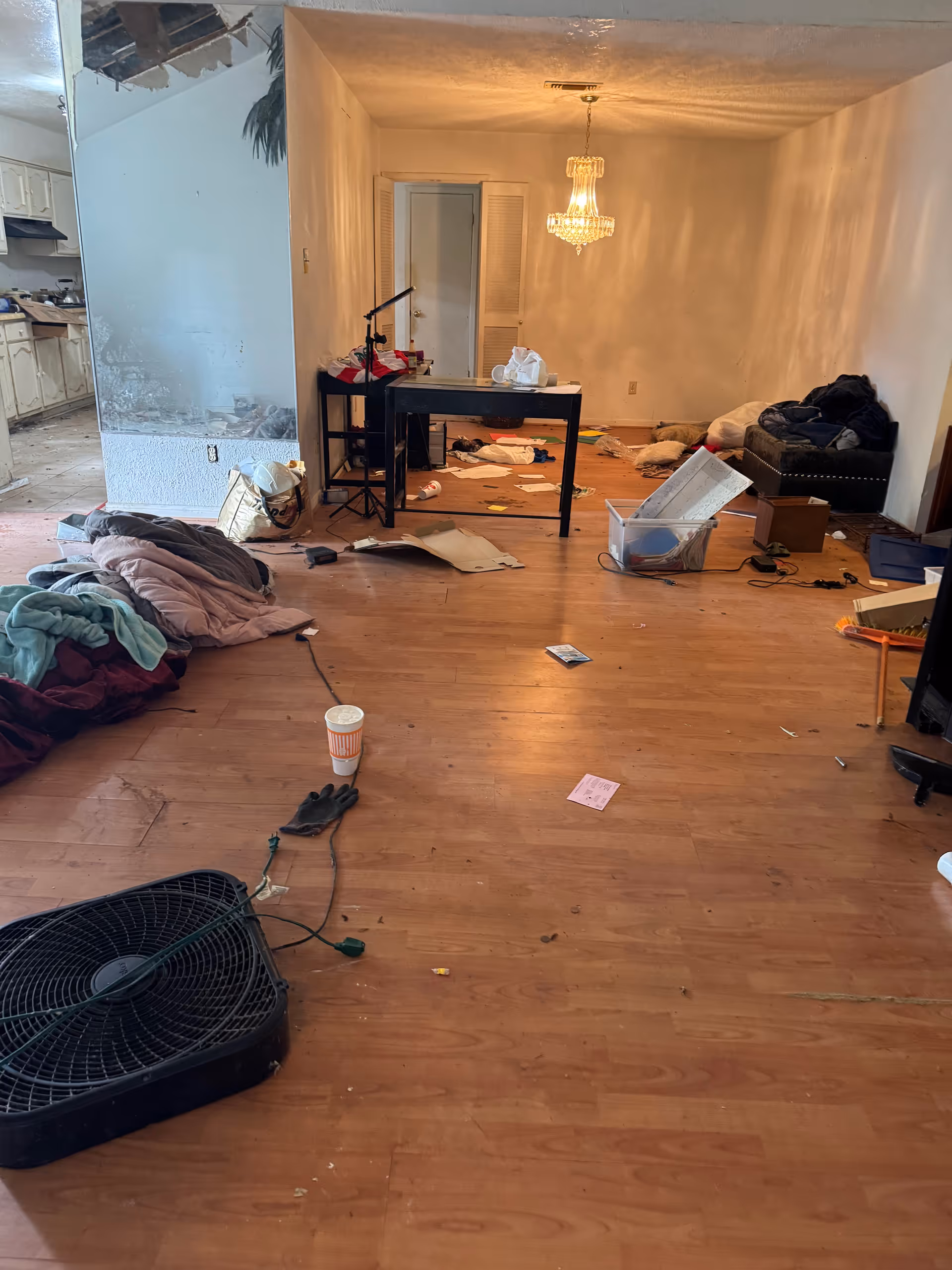 Messy living room with scattered papers, clothes, a fan, coffee cup, and cluttered furniture.