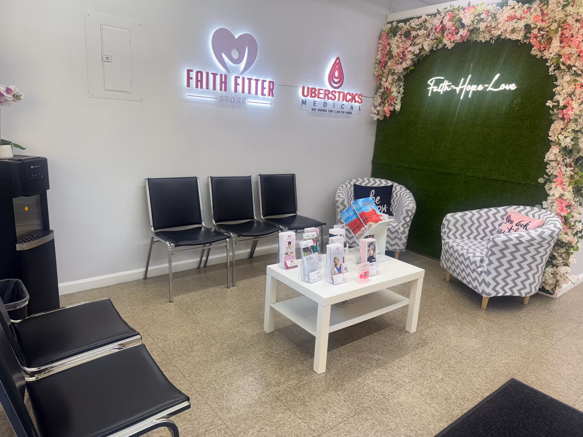 Interior waiting area with black chairs, a white coffee table, brochures, and a floral green wall.