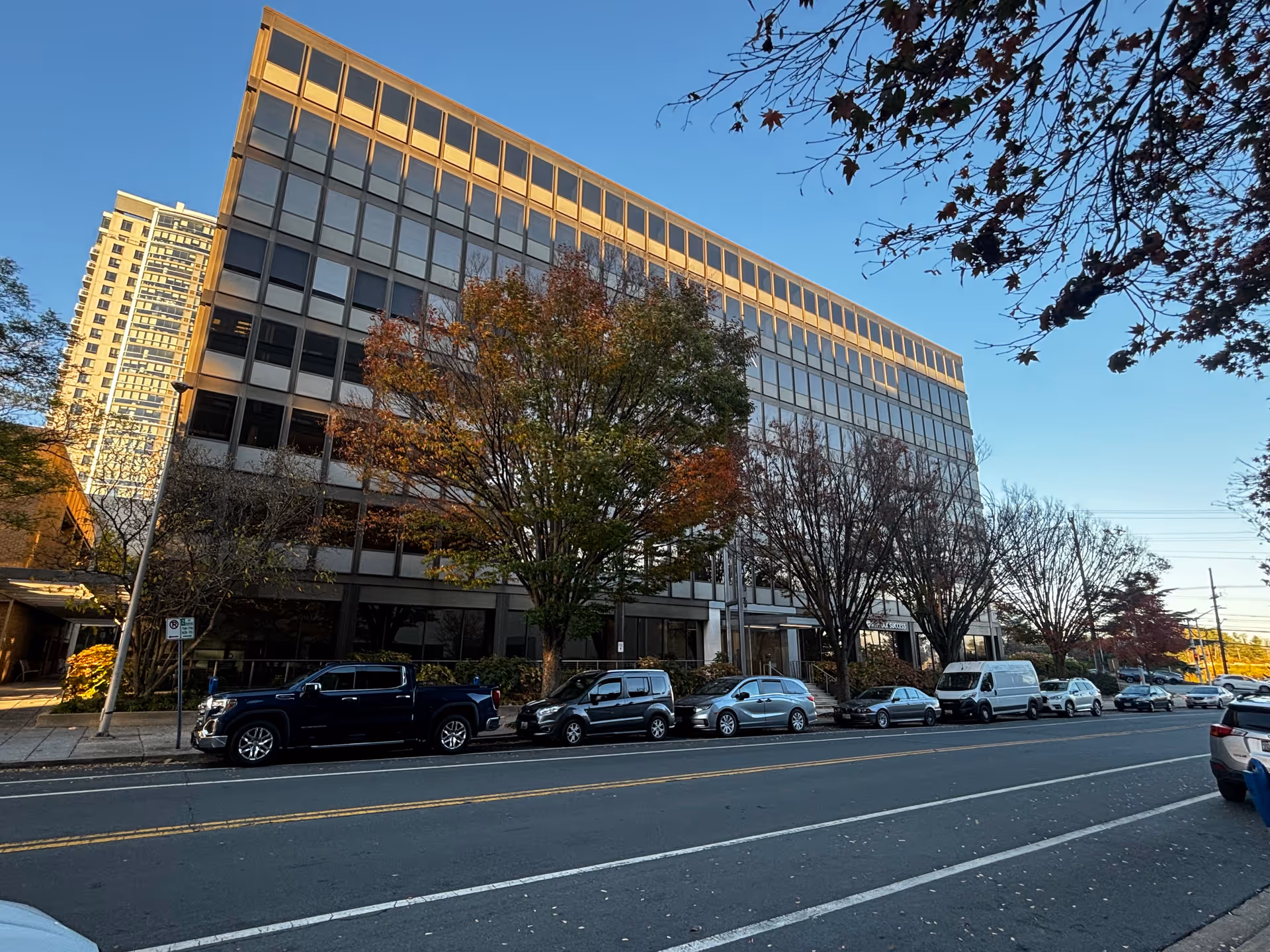Street view of a multi-story office building with reflective windows, several parked cars along the street, and trees with autumn foliage in front of the building under a clear blue sky.
