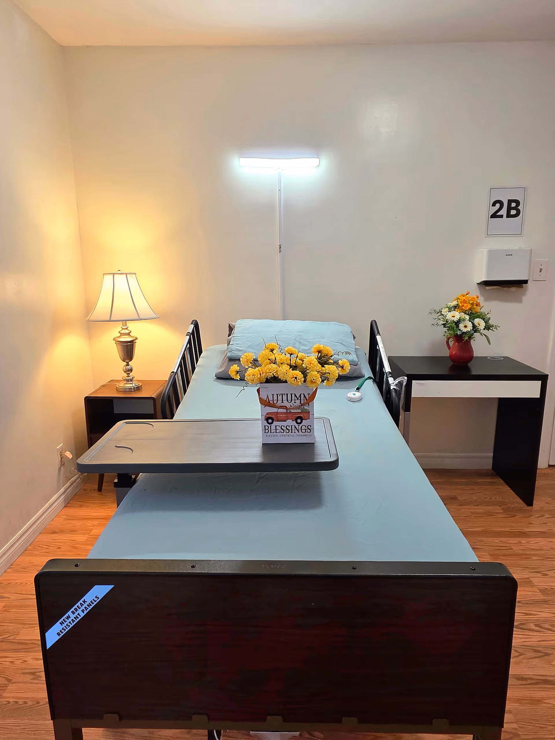 Single patient bed in a small room with a lamp, nightstand, flowers, and overbed tray.
