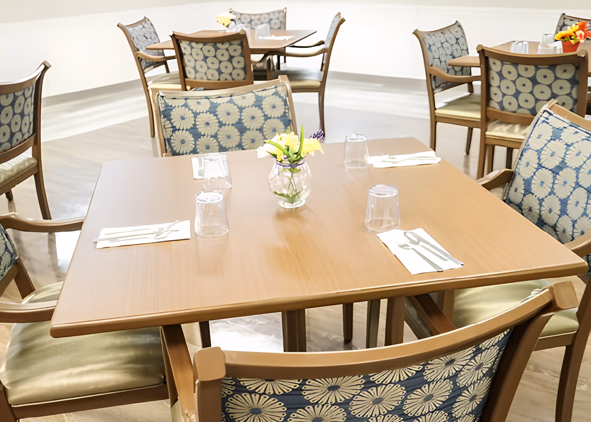 Empty dining room with wooden tables and patterned chairs set with napkins, utensils, plastic cups, and small flower vases.