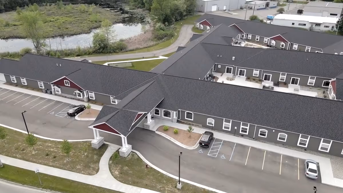 Aerial view of Hampton Manor of Brighton Premier Assisted Living & Memory Care building with a covered entrance, parking spaces including handicapped spots, surrounding greenery, and a pond in the background.