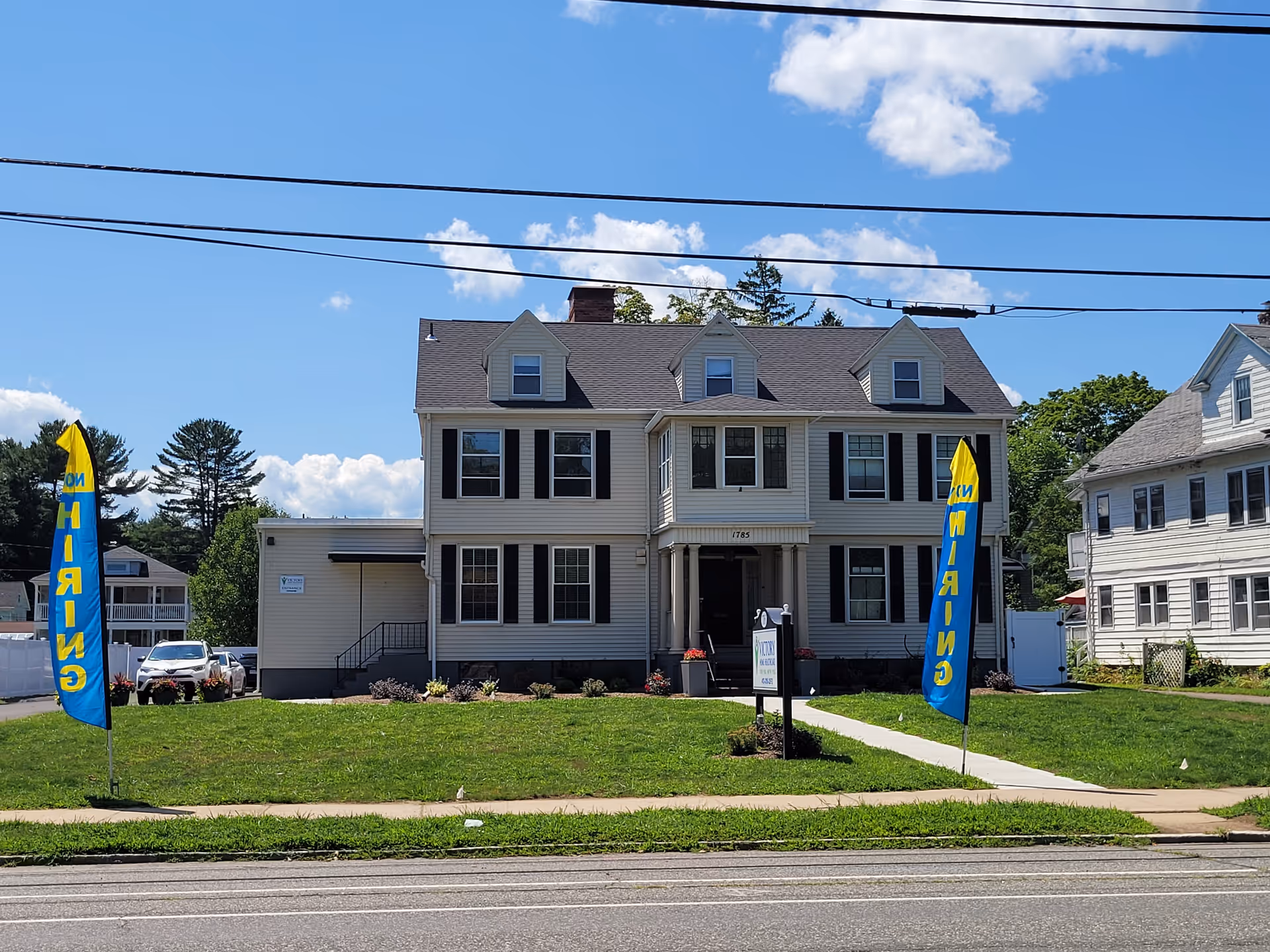 Two-story beige building with black shutters, blue banners, and a manicured front lawn.