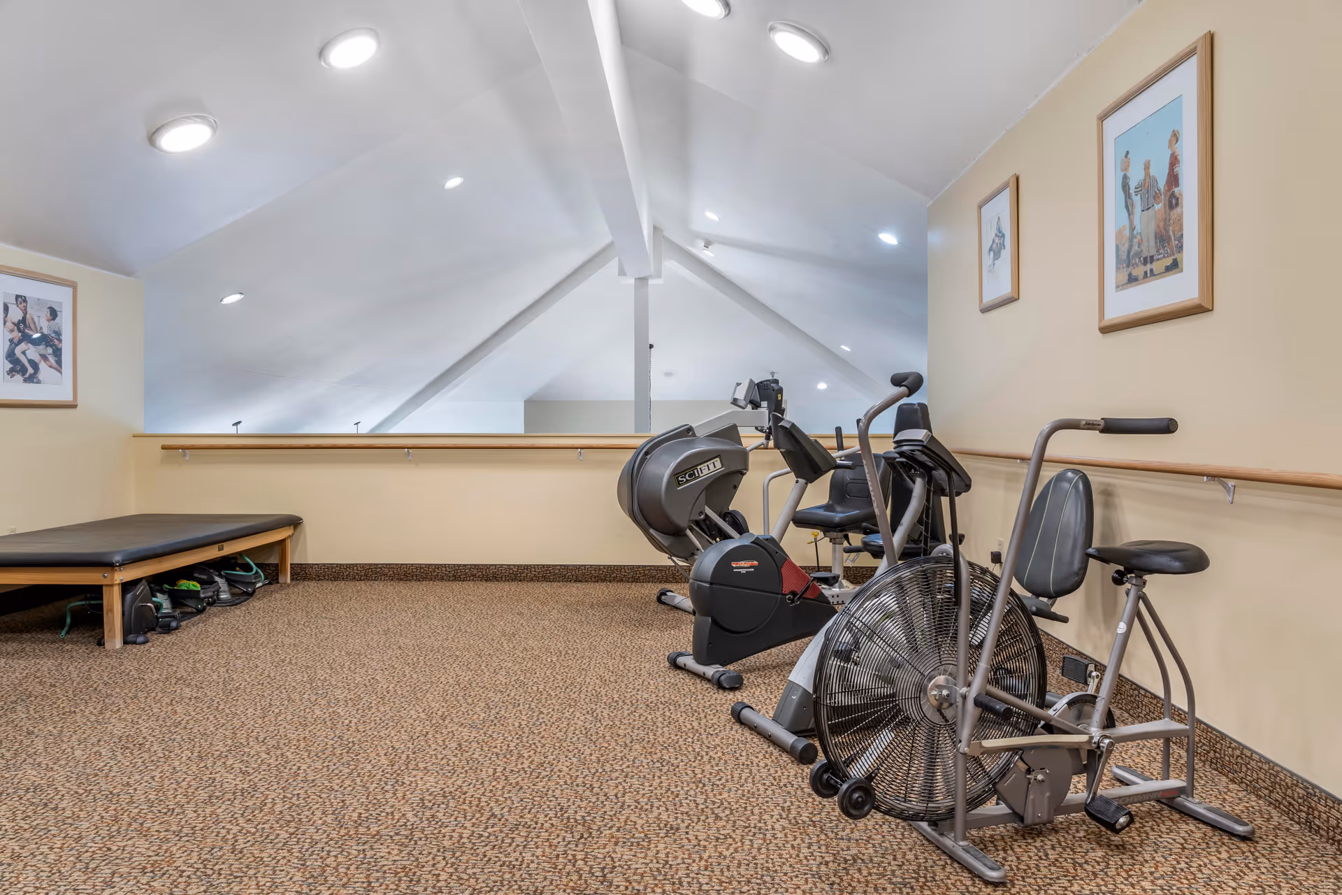 A small exercise room with beige walls and carpeted floor featuring three pieces of exercise equipment including a stationary bike, a recumbent bike, and an air resistance bike. There is a padded bench with exercise bands underneath it on the left side and framed artwork hanging on the walls.