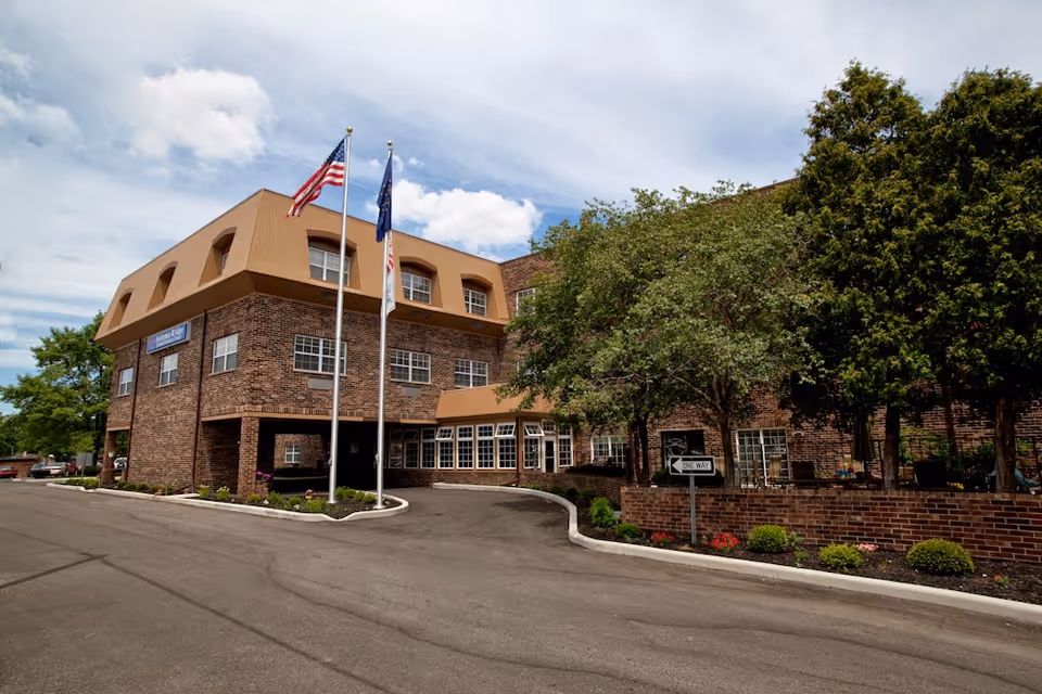 Exterior view of a multi-story brick building with a brown mansard roof, two flagpoles with flags, and landscaped greenery including trees and shrubs. The building has multiple windows and a driveway with a one-way sign.