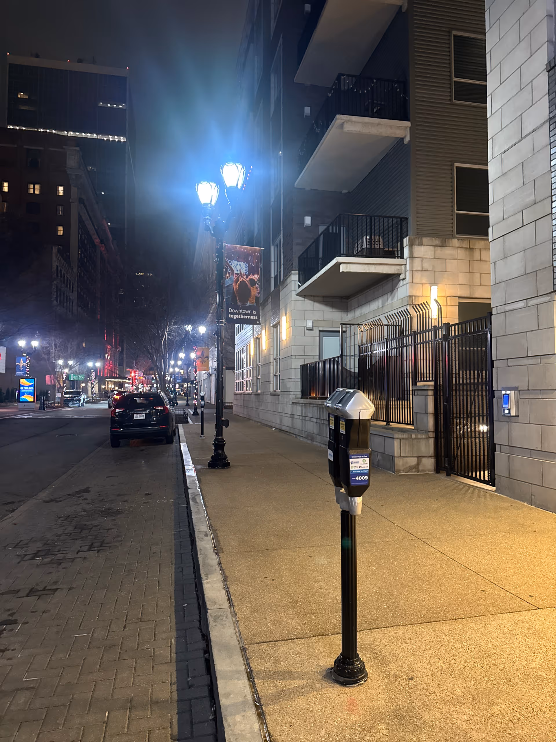 Nighttime city street with a lit sidewalk, parking meter, and modern building exterior.