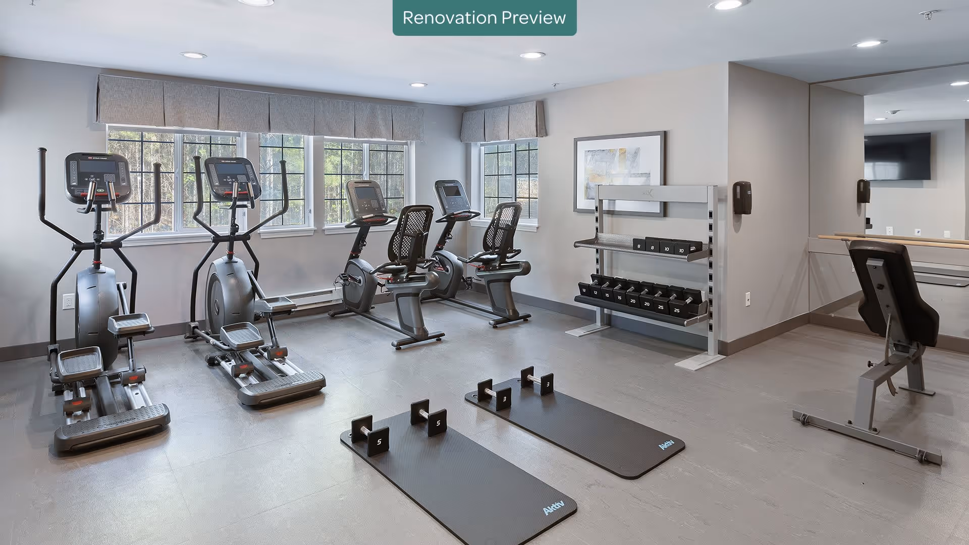 A modern fitness room with exercise equipment including elliptical machines, stationary bikes, a weight bench, dumbbells on a rack, and two exercise mats with small weights. Large windows let in natural light, and a mirror covers one wall. The room has a clean, neutral color scheme with gray floors and light walls.