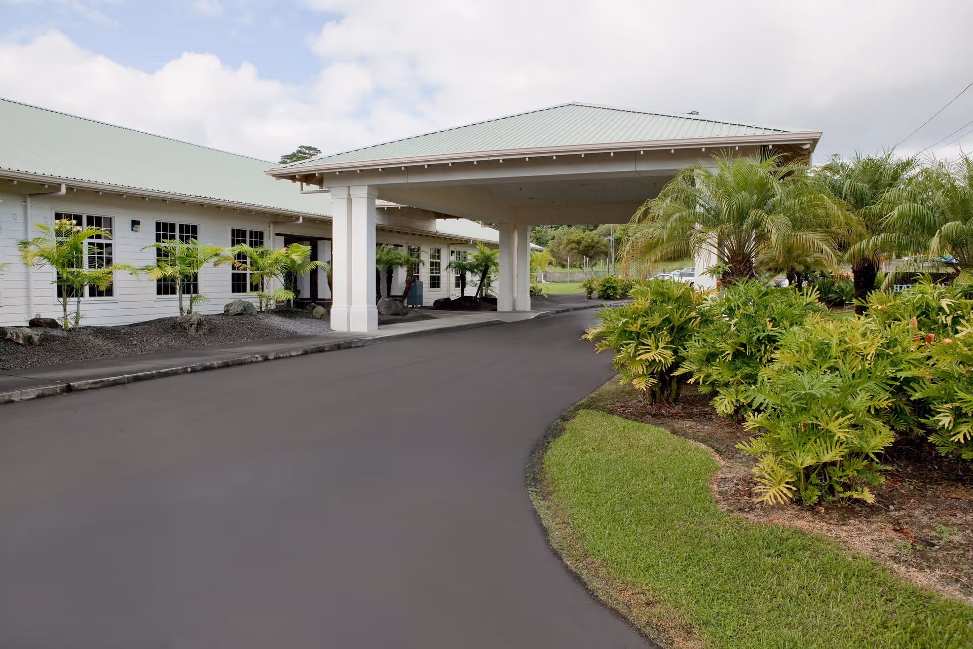 Light-colored, single-story building with a covered entrance and tropical plants along a paved driveway.