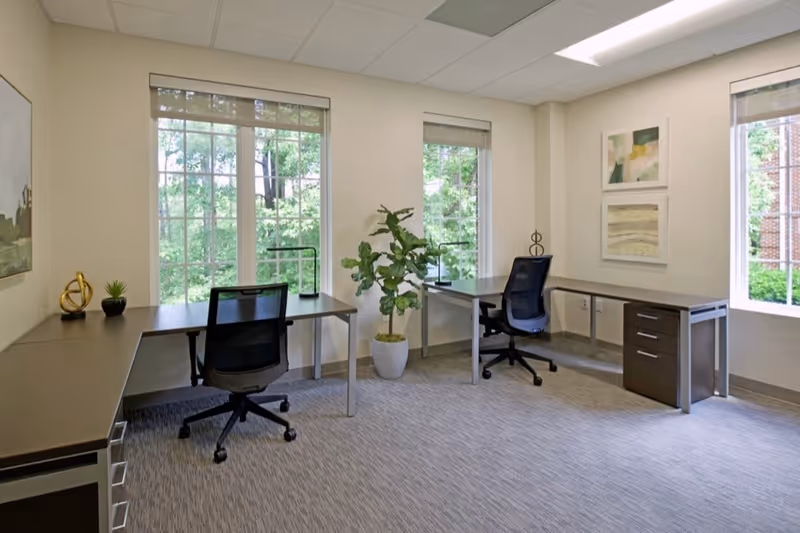 Interior office area with two desks, chairs, a plant, and windows.