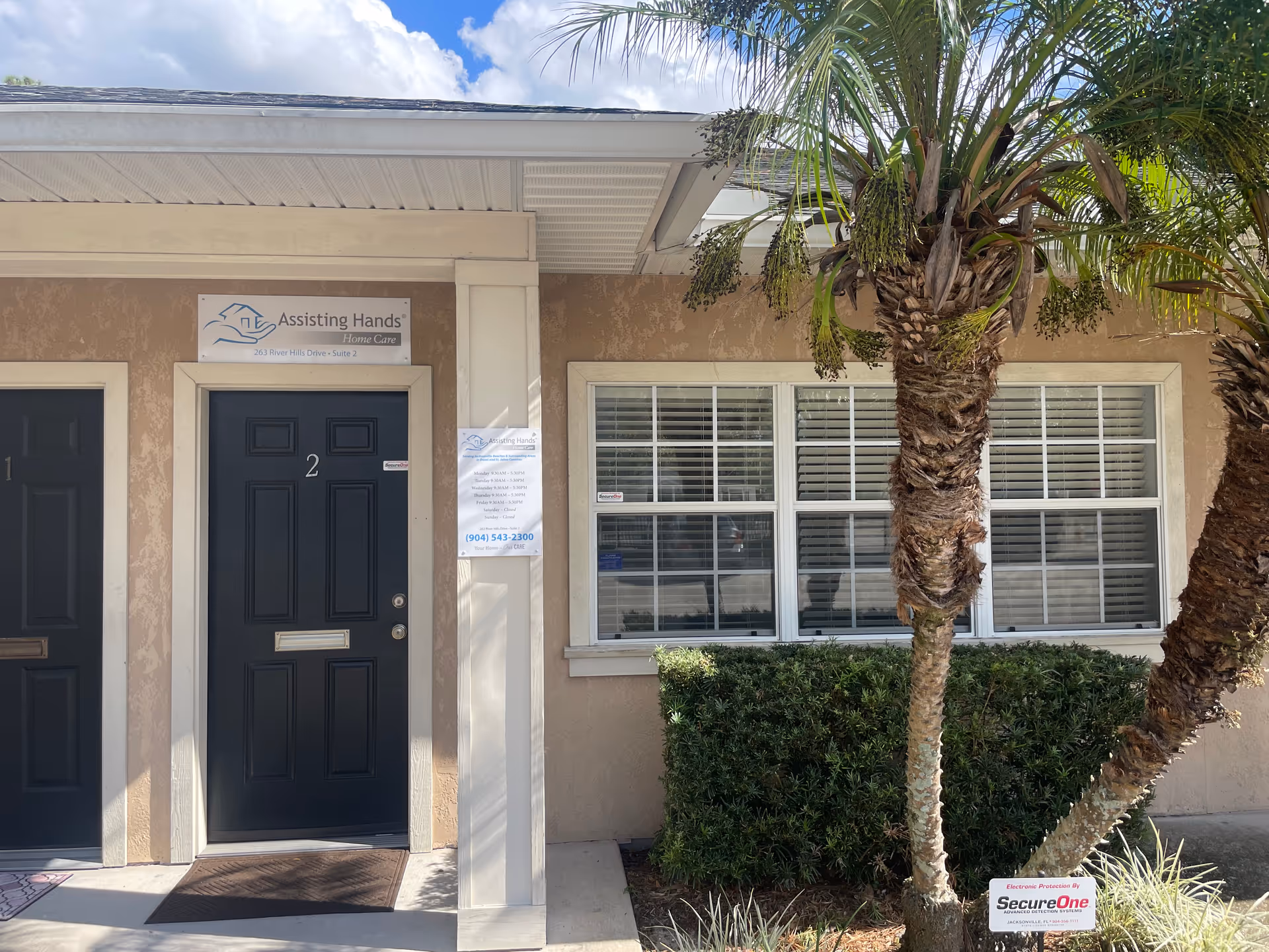Exterior of Assisting Hands Home Care facility with doors, windows, palm trees, and hedges.