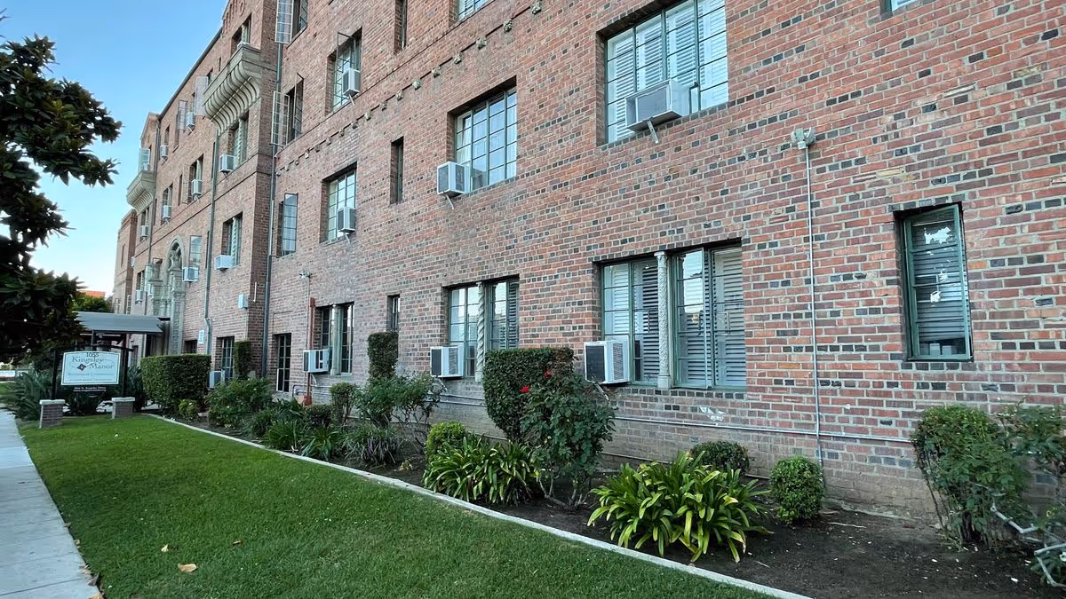 Front exterior of Kingsley Manor, a brick multi-story building with windows, window air conditioners, landscaped shrubs and a lawn by the sidewalk.