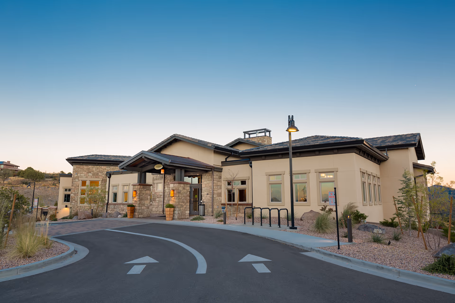 Front exterior of a single-story community building with a covered stone and stucco entrance, driveway arrows, lamp posts and landscaped grounds.