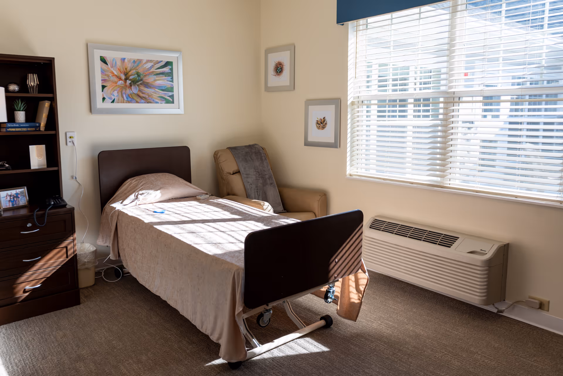 A well-lit bedroom in a senior living facility with a single hospital-style bed covered with a beige blanket. Next to the bed is a comfortable armchair with a gray throw draped over it. The room has a large window with white blinds allowing sunlight to stream in. On the left side, there is a dark wooden dresser with shelves holding books, a small plant, and a framed photo. The walls are decorated with three framed pictures, including a large floral artwork above the bed.