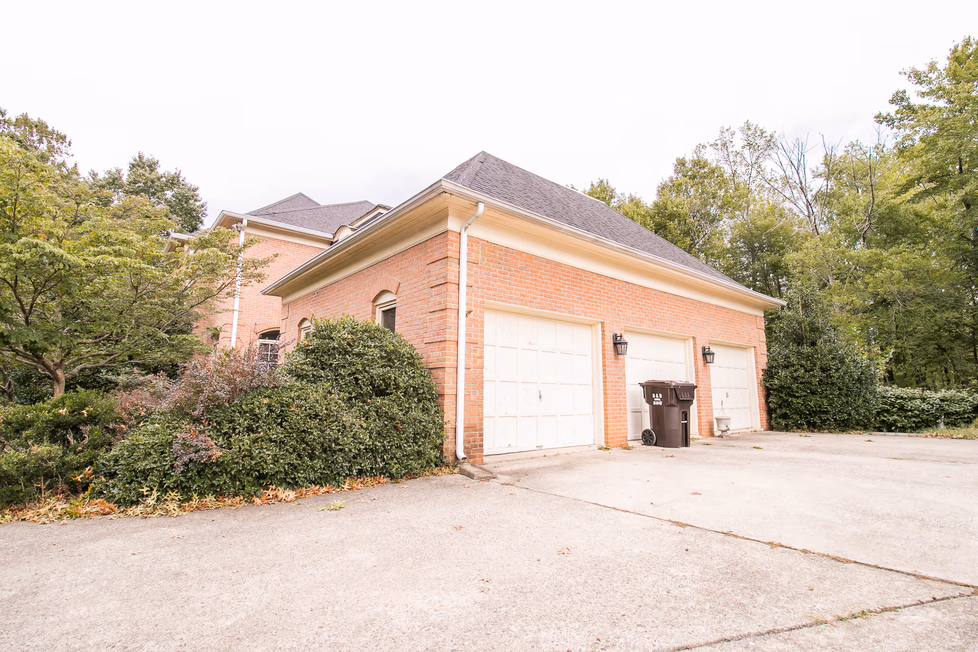 Exterior view of a brick building with three white garage doors and a driveway in front. The building is surrounded by green trees and bushes under a cloudy sky.