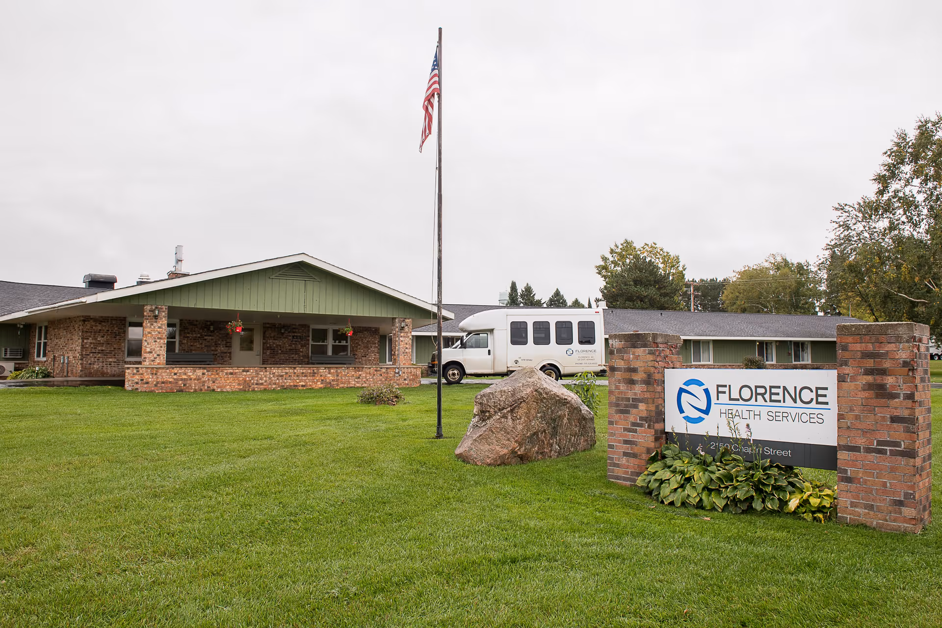 Exterior view of Florence Health Services building with a green lawn, an American flag on a flagpole, a large rock, and a sign displaying the facility's name. A white shuttle bus is parked near the building.