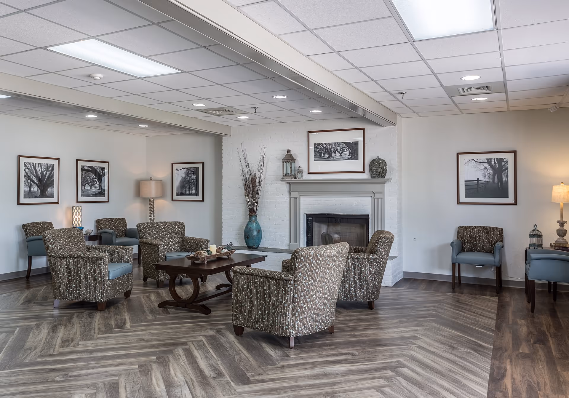 A cozy senior living facility lounge area with patterned armchairs arranged around a wooden coffee table. The room features a white brick fireplace with decorative items on the mantel, framed black and white tree photographs on the walls, and soft lighting from table lamps. The floor has a wood-patterned design and the ceiling has recessed lighting and a drop ceiling.