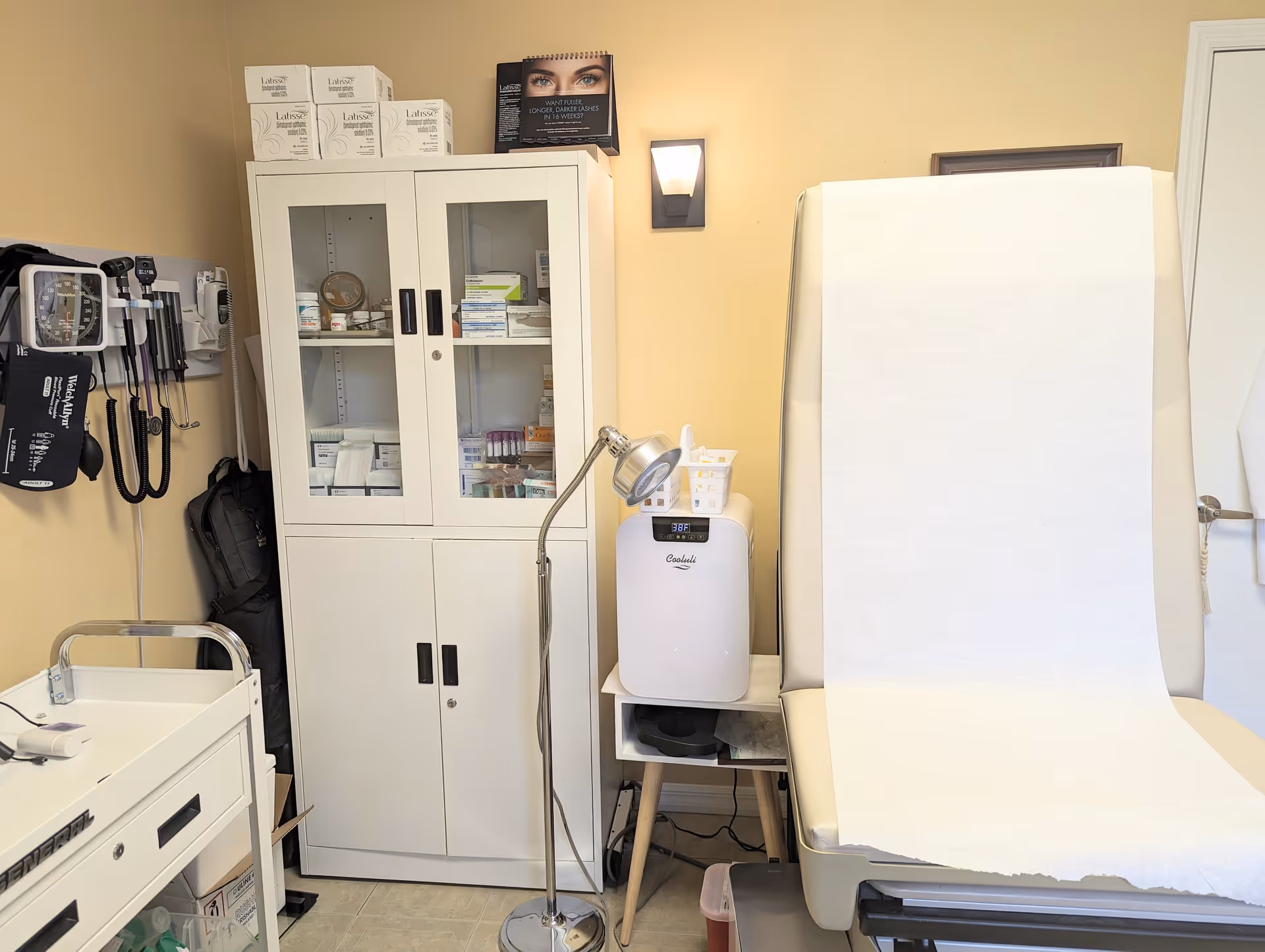 Medical examination room with a white cabinet, exam bed, and assorted medical equipment.
