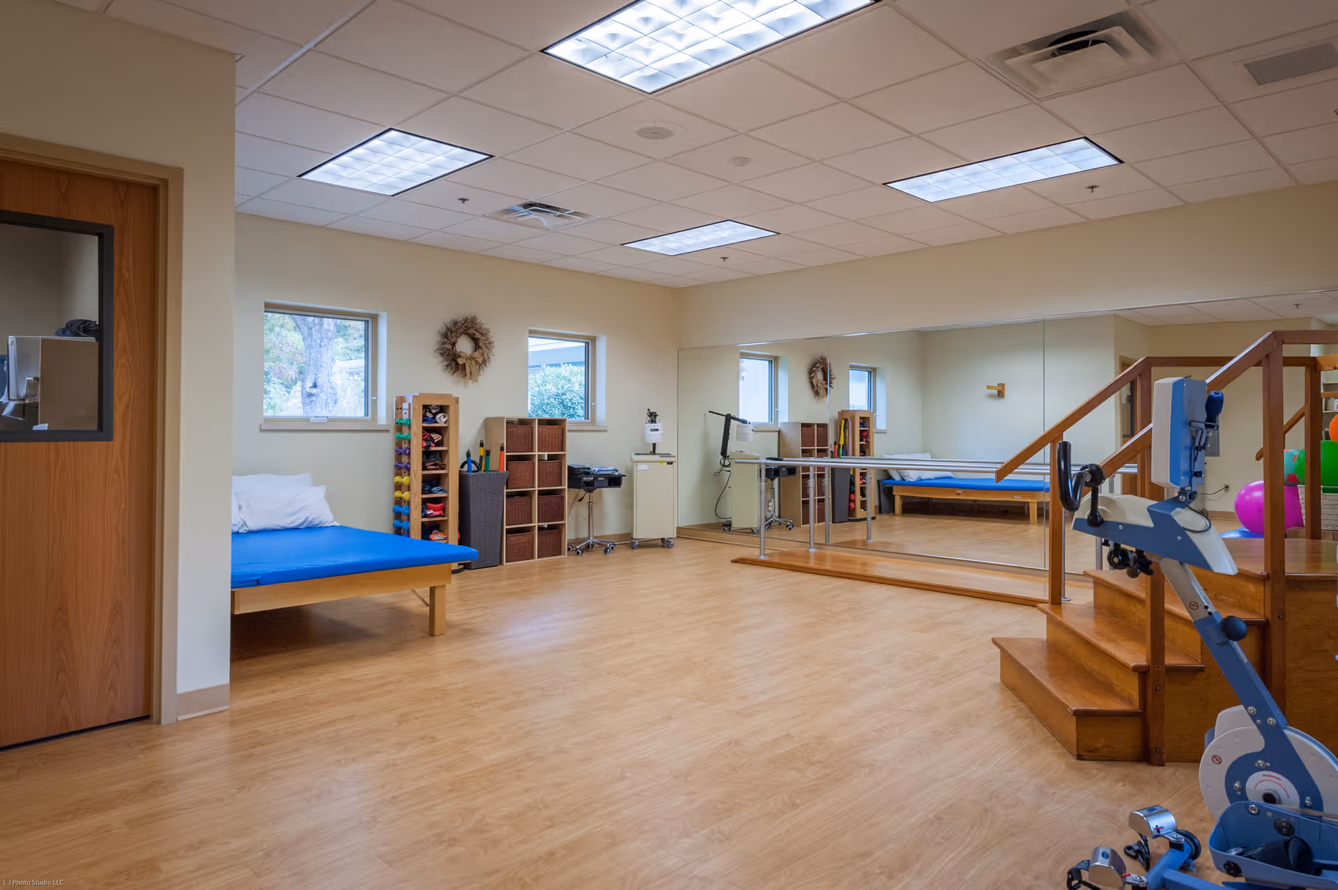 A spacious therapy or exercise room with wooden flooring, a blue padded therapy table with pillows, a set of stairs with handrails, exercise equipment including a stationary bike, large wall mirrors, and shelves with exercise tools and colorful weights. The room has beige walls, two small windows, and a wooden door with a window.