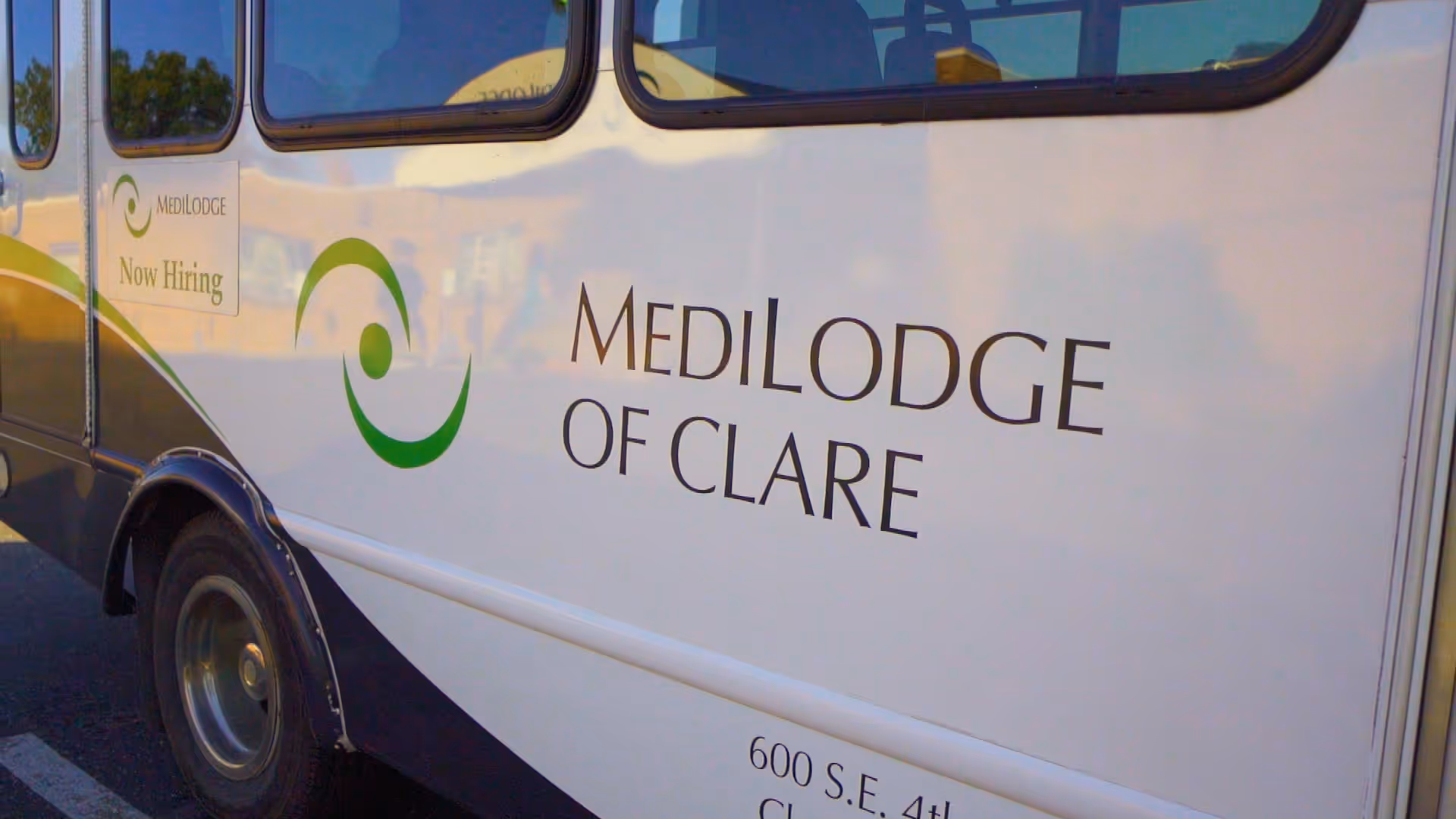 Side view of a white and green shuttle bus with the text 'Medilodge of Clare' and 'Now Hiring' displayed on the side.
