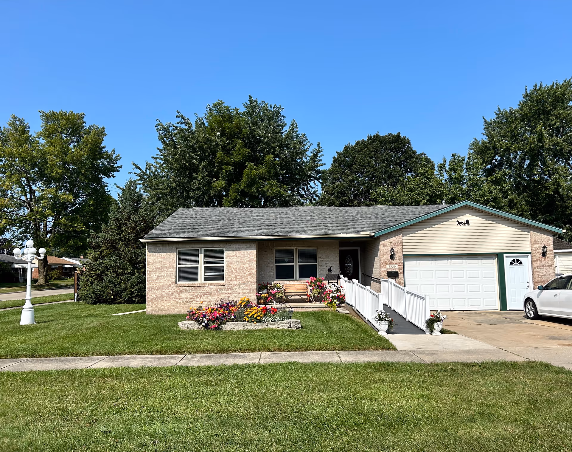 Single-story brick house with a gray roof, a white garage door, and a white accessibility ramp leading to the front door. There is a flower bed with colorful flowers and a bench in front of the house. A white car is parked in the driveway, and a white lamp post is on the lawn. Trees and a clear blue sky are visible in the background.