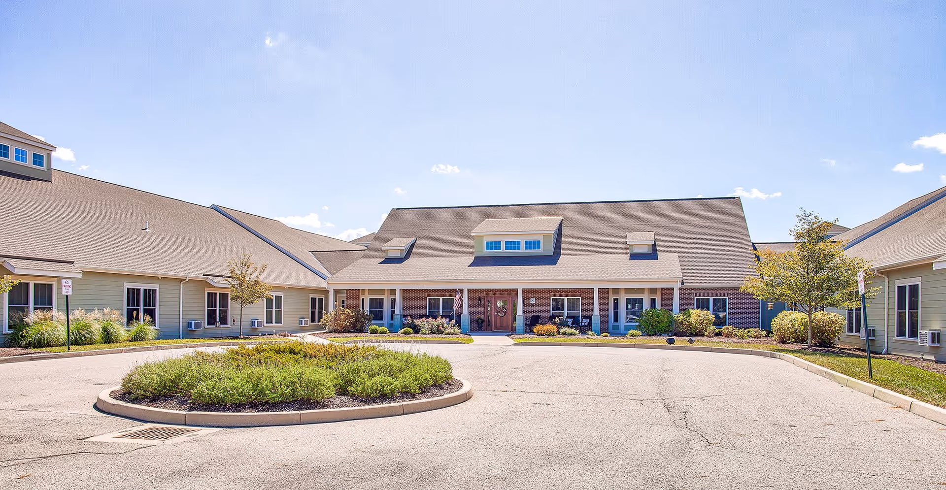 Front exterior view of Villages of Huber facility showing a large building with a peaked roof, multiple windows, and a central entrance with a covered porch. There is a circular driveway with landscaped greenery in the center and small trees and shrubs around the building under a clear blue sky.