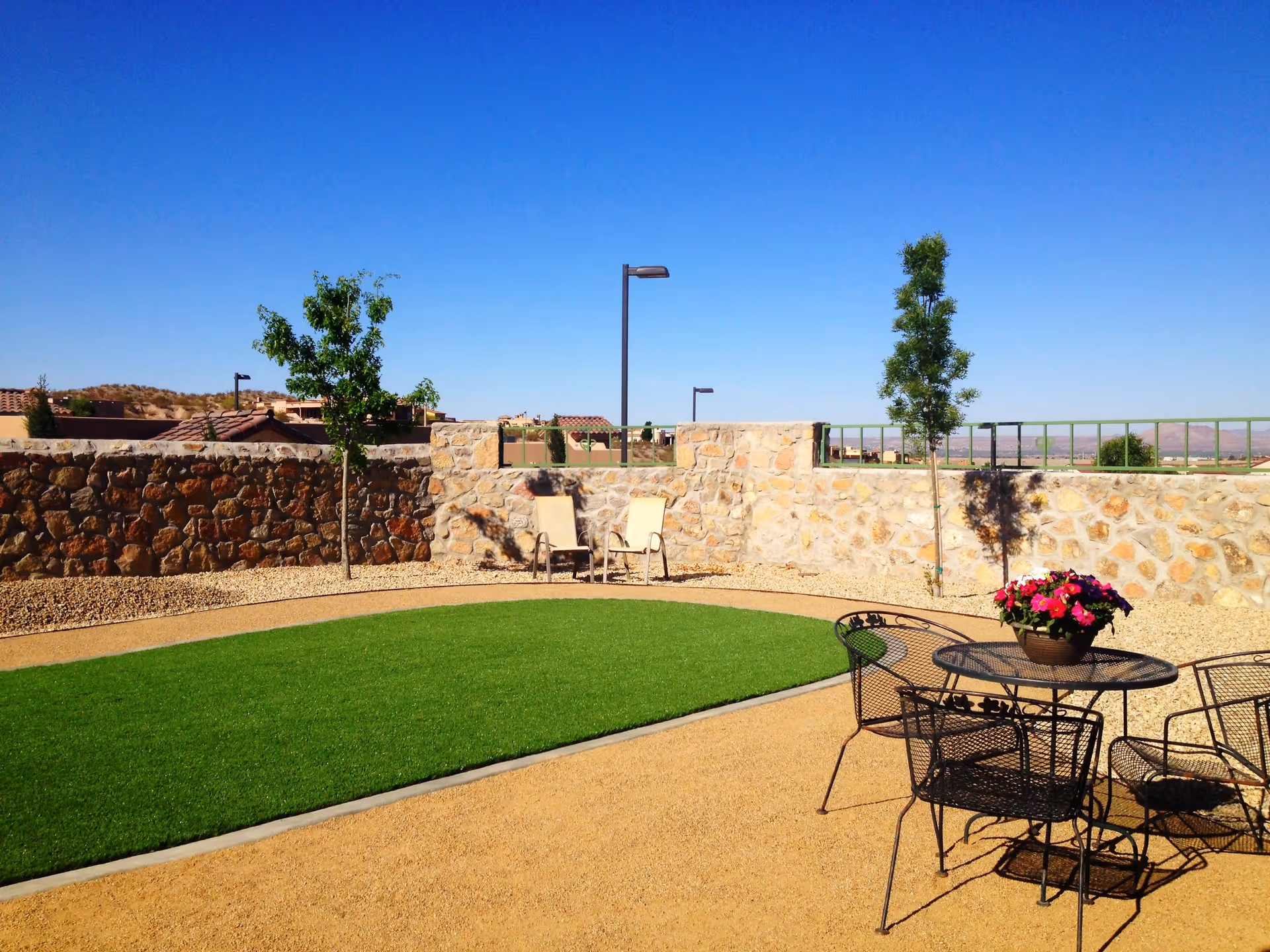 Outdoor patio area with a small green lawn surrounded by gravel, two young trees, a stone wall, two beige lounge chairs, and a black metal table with four chairs and a flower pot with pink flowers on the table under a clear blue sky.