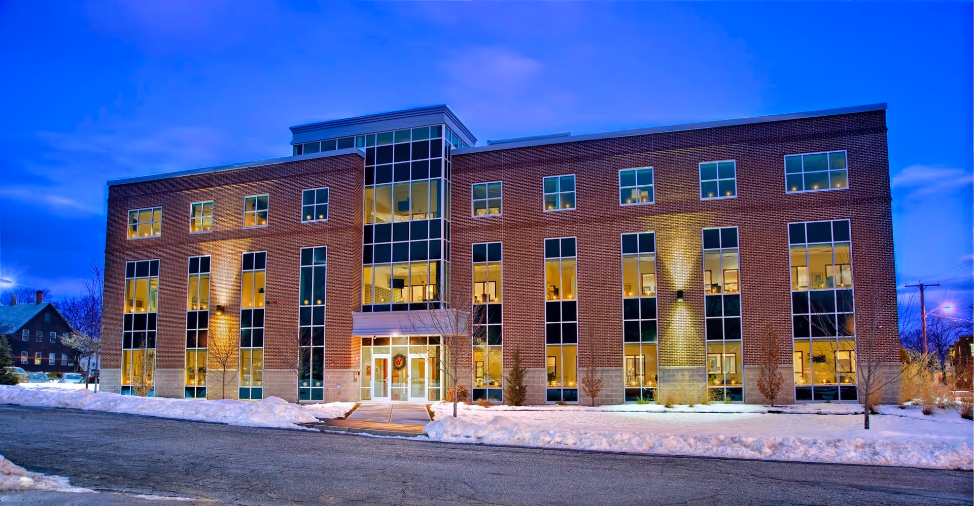 Three-story brick senior living building with large glass windows and a snowy sidewalk.