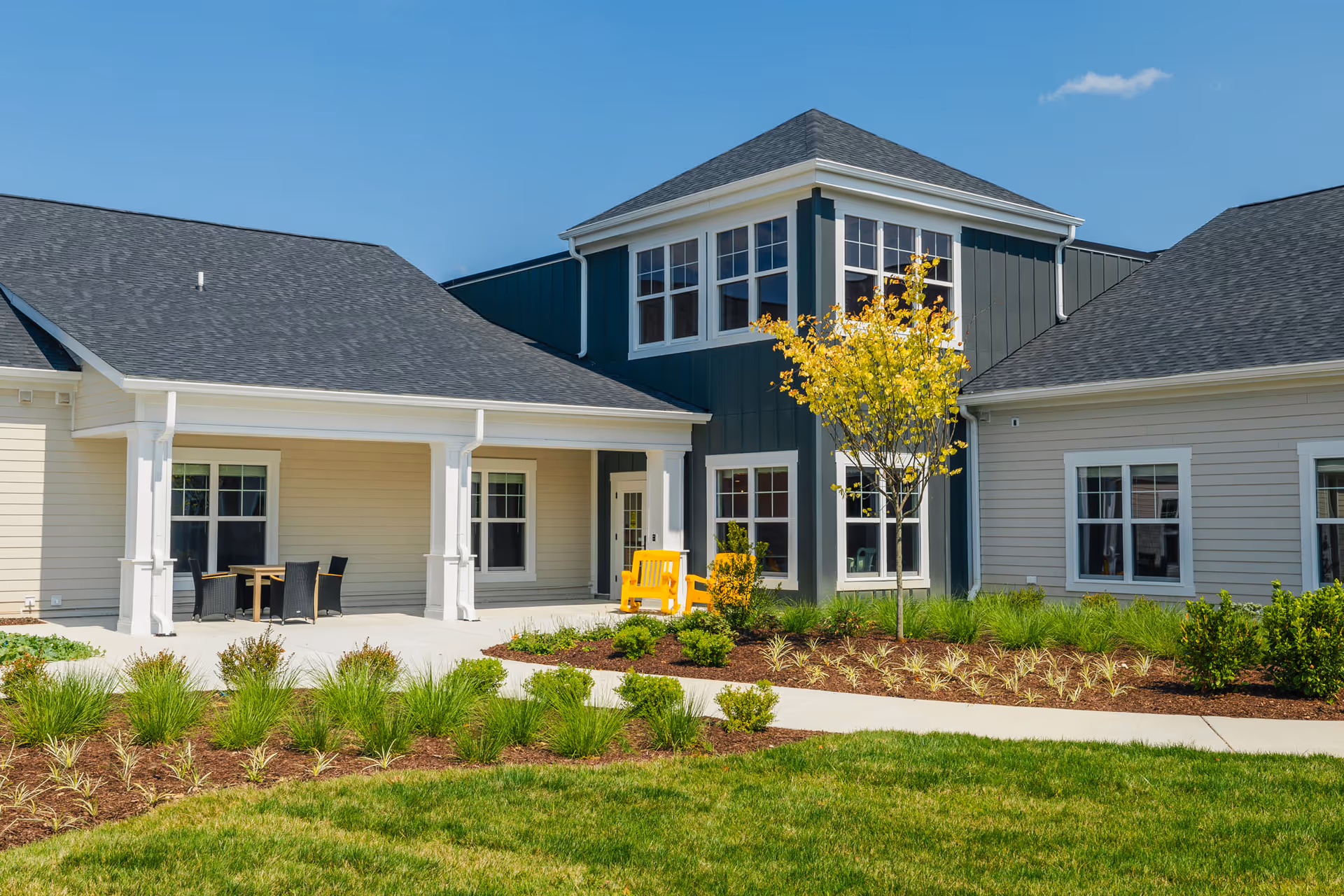 Exterior view of a senior living facility building with beige and dark green siding, multiple windows, a small tree with yellow leaves, landscaped garden beds, a concrete walkway, and outdoor seating including a yellow bench and a table with chairs under a covered porch.