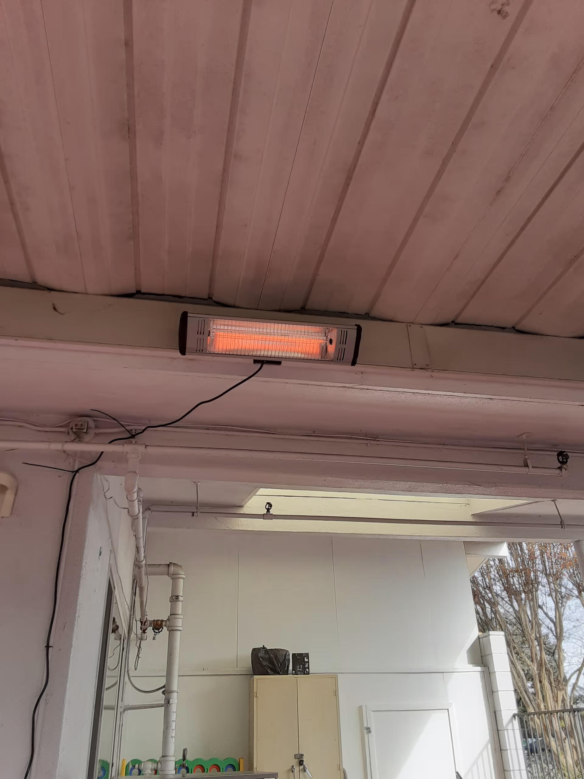 Outdoor covered area with a wall heater glowing, exposed pipes, and white walls.