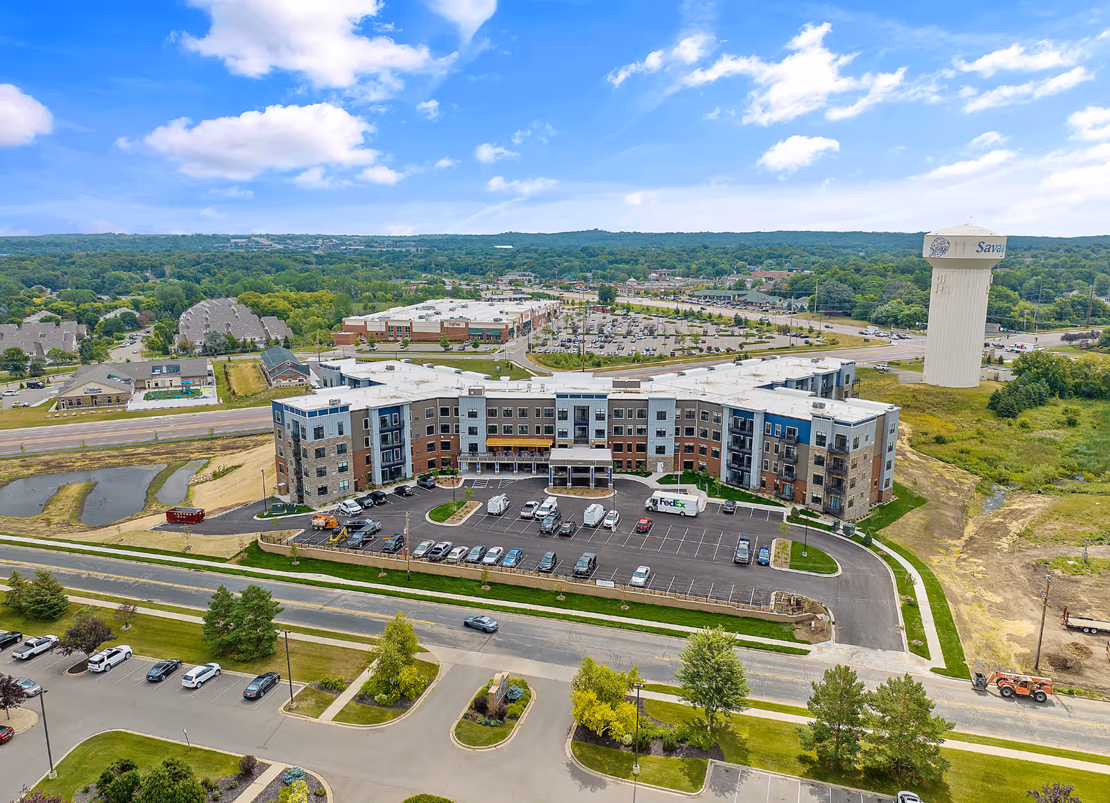Aerial view of a four-story senior living building with a front parking lot, surrounding roads, and a nearby water tower.