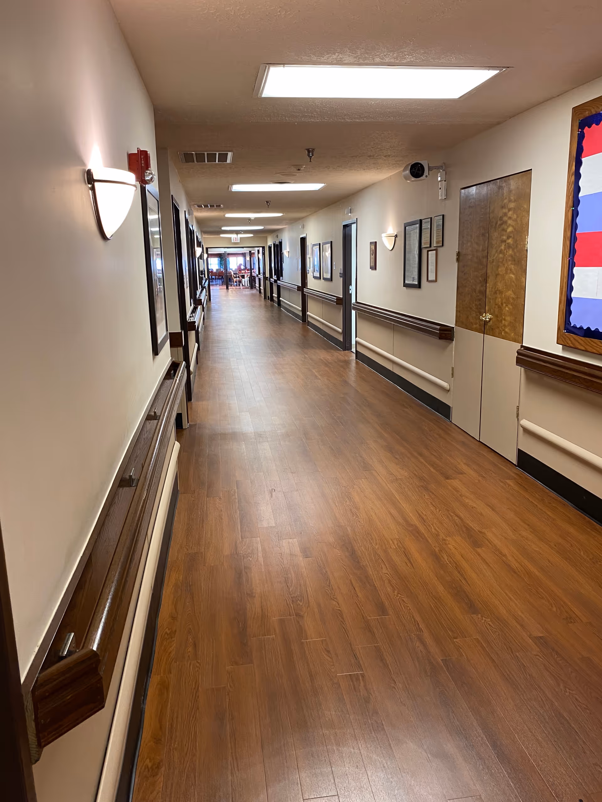 Long interior hallway in a senior living center with wood flooring, doors, and sconces.