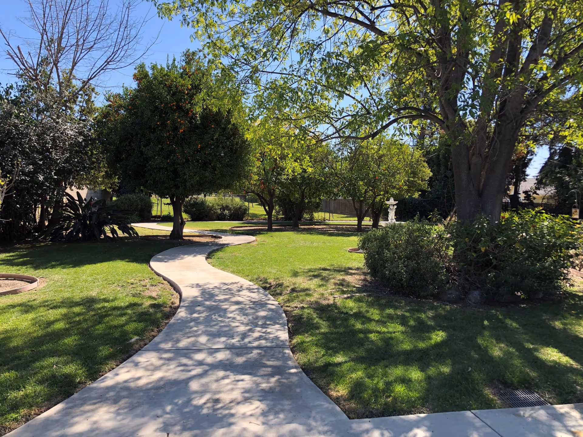 Curving concrete walkway through a shaded garden with trees, lawn, and a small statue in the background.