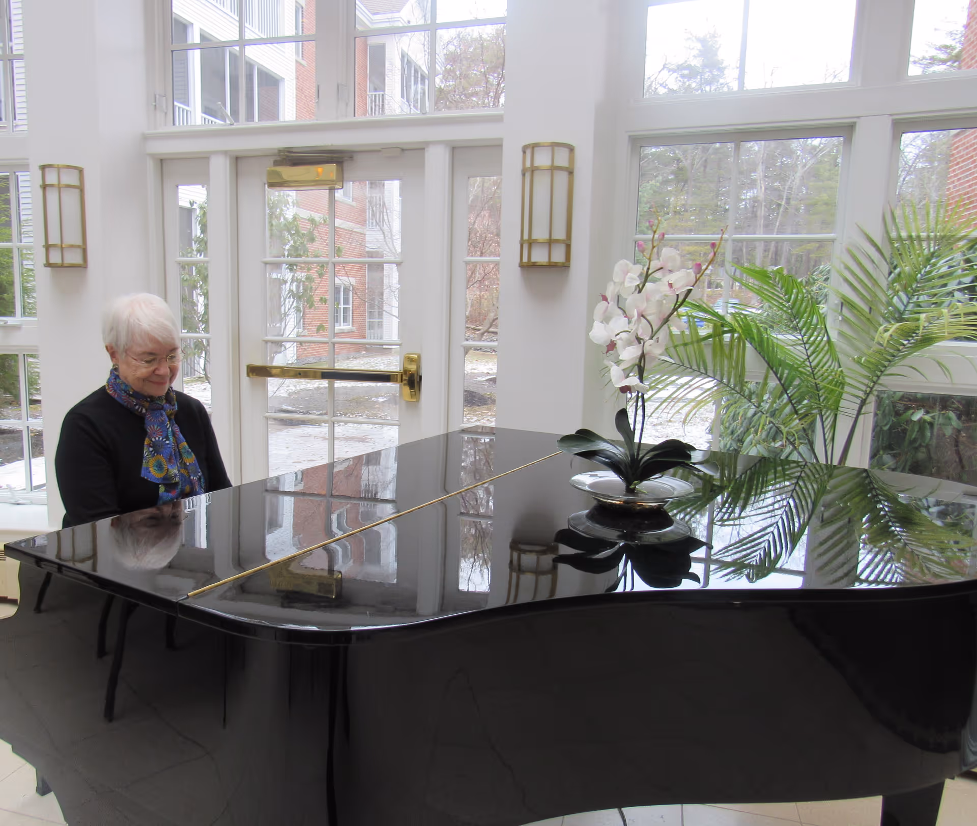 An elderly woman with white hair and glasses is sitting at a black grand piano in a bright room with large windows. The piano has a decorative white orchid and green plant on top. Outside the windows, trees and a brick building are visible.