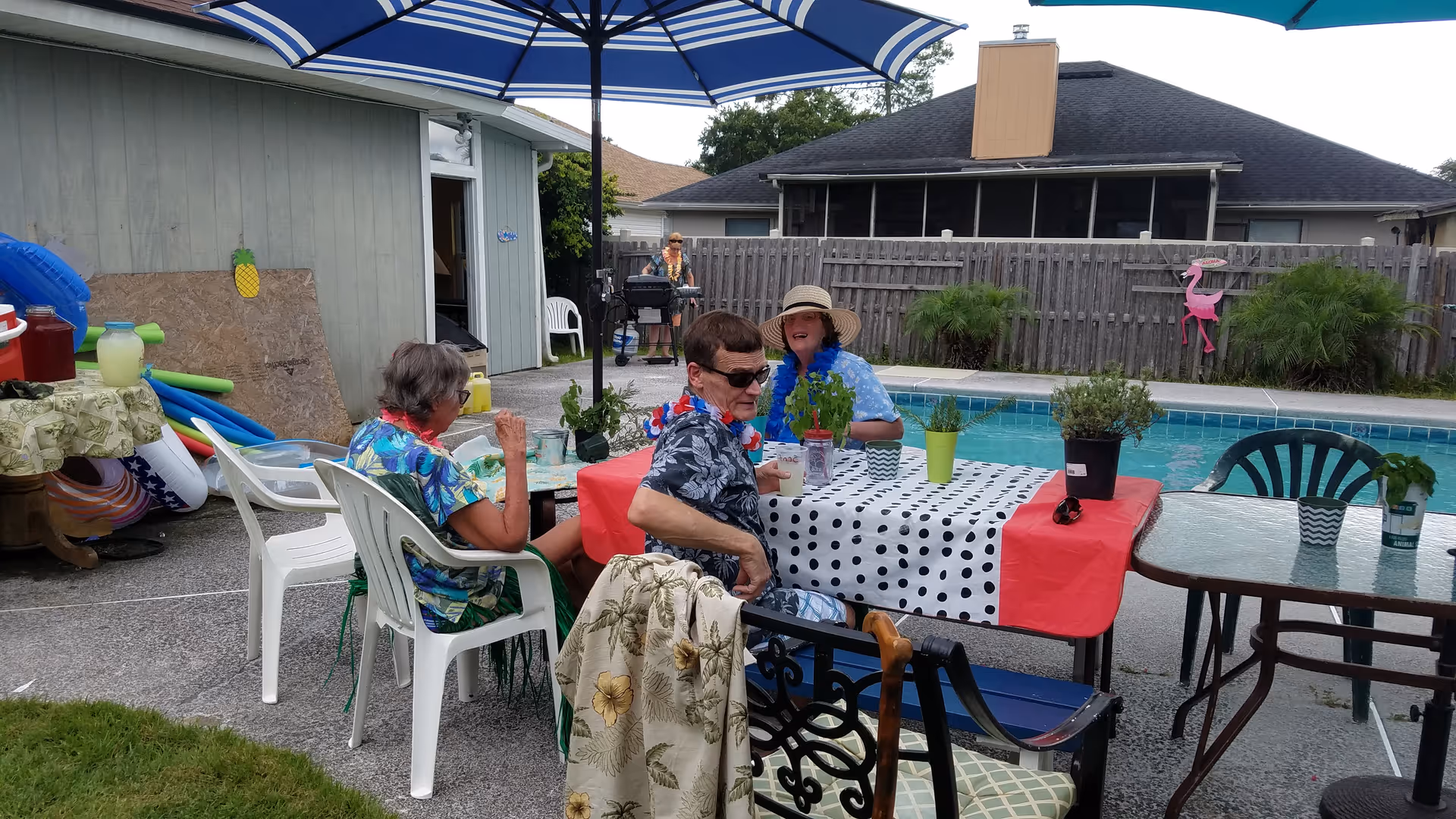 Elderly residents sit at a poolside table under umbrellas, enjoying a sunny outdoor gathering.