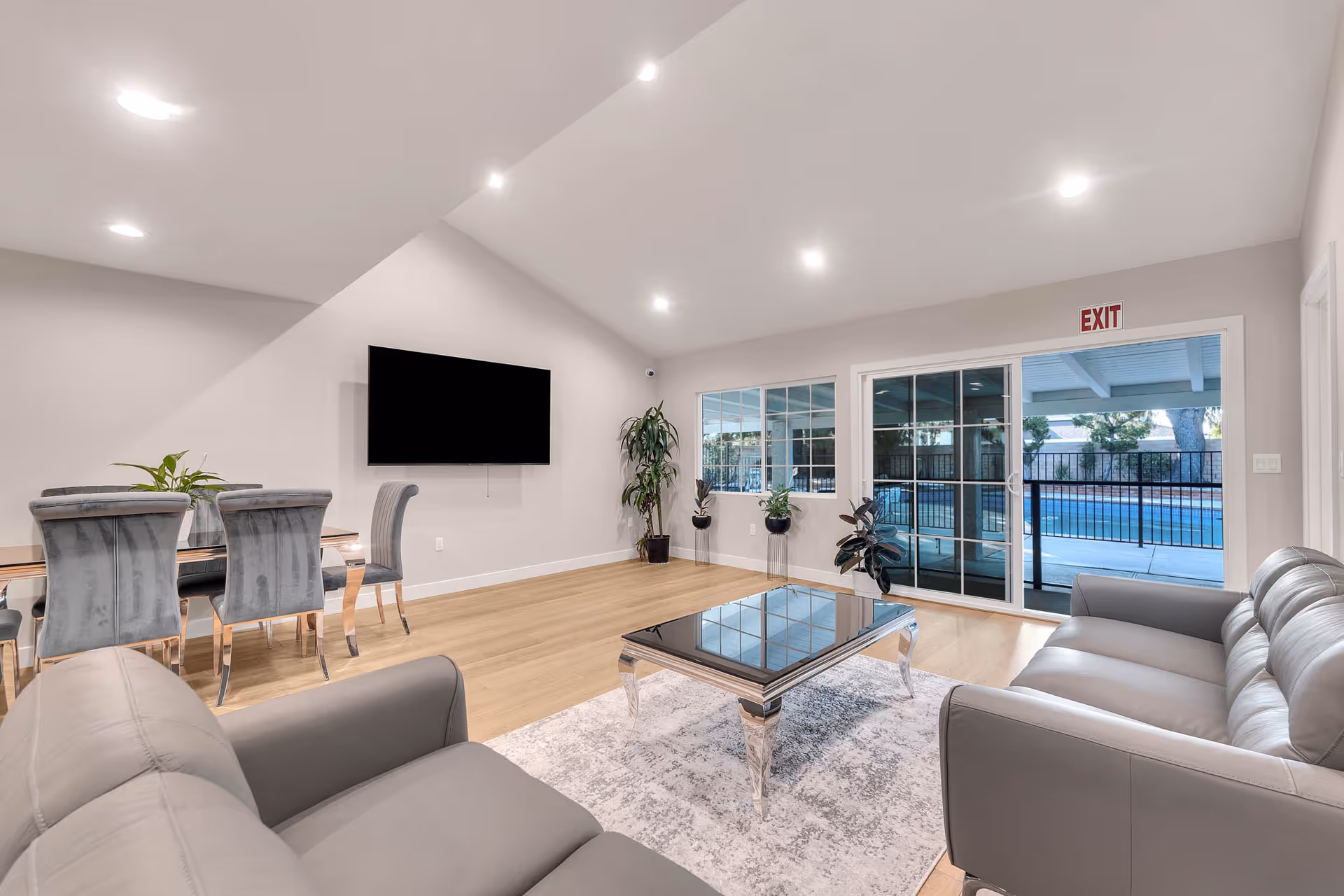 Bright modern senior living lounge with couches, dining area, TV, plants, and glass doors.