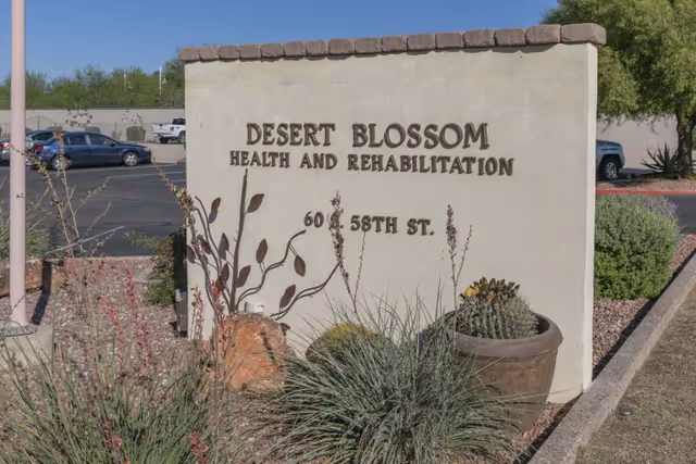 Entrance stone sign reading "Desert Blossom Health and Rehabilitation" surrounded by desert plants and a parking lot in the background.