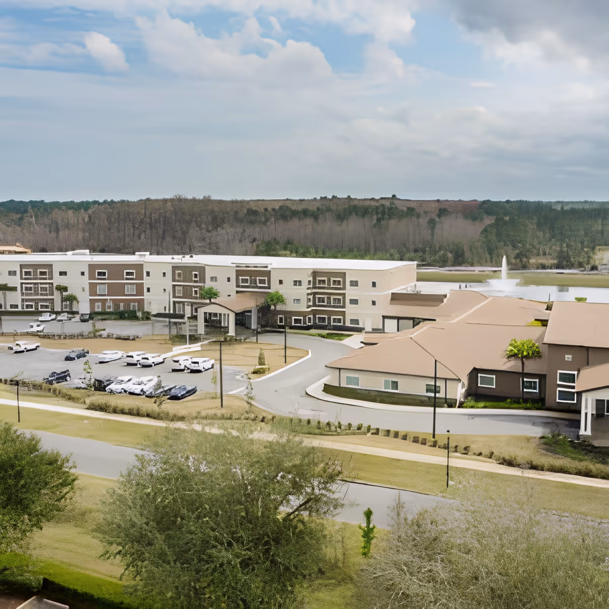 Aerial view of Discovery Village At Deerwood Assisted Living & Memory Care facility showing multiple connected buildings with beige and brown exteriors, a parking lot with several cars, landscaped greenery, a road in the foreground, and a water fountain in a pond behind the buildings under a partly cloudy sky.