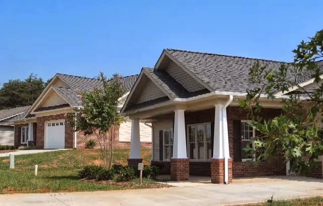 Single-story brick building with a covered porch supported by white columns, driveway, and small landscaped lawn under a clear blue sky.
