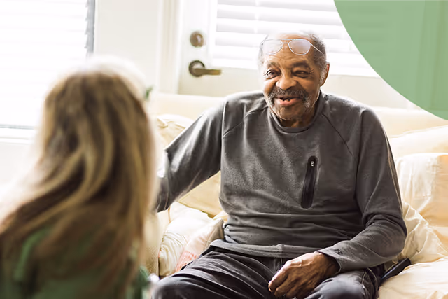 Smiling elderly man on a beige sofa chats with a caregiver indoors in a living room.
